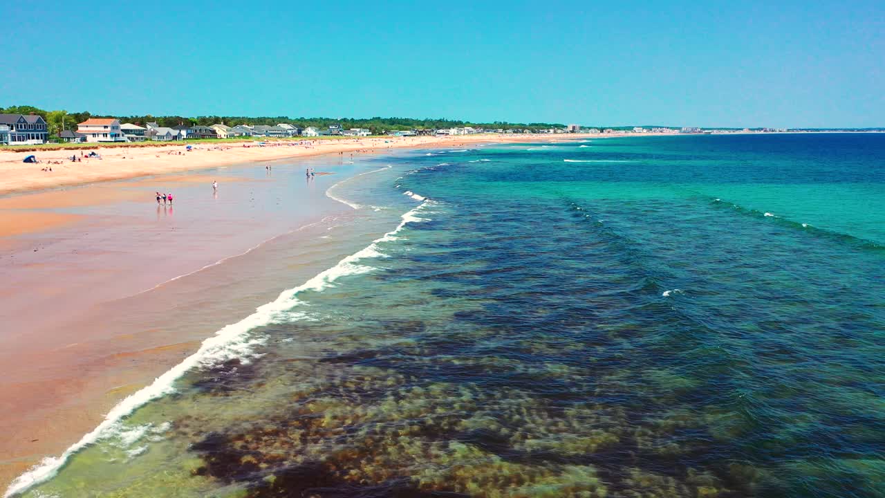 Drone view shows crowds on Saco, Maine’s beaches. Tourists relax, kids splash in waves, houses stand along the shore. Peak-season East Coast vibe with ocean, sand, and coastal New England charm