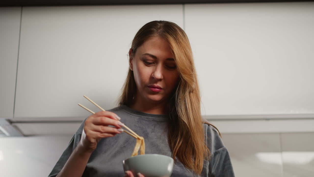 Young woman with long flowing hair stands in bright modern kitchen eating noodles with chopsticks while looking thoughtfully into background