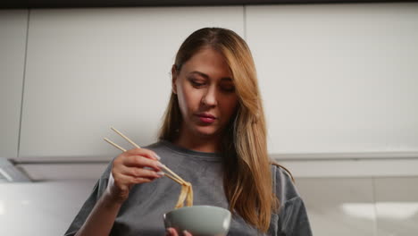 young woman with long flowing hair stands in bright modern kitchen eating noodles with chopsticks while looking thoughtfully into background
