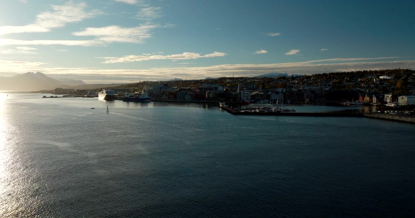 Panoramica aerea del porto di Tromso sotto il basso sole autunnale con cime innevate e acqua calma, carrellata panoramica con drone verso la silhouette della città