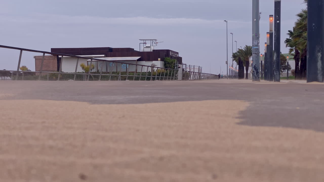 Passeio marítimo de Sanlúcar de Barrameda em um dia ventoso, com areia sendo soprada pelo caminho perto de edifícios costeiros e postes de iluminação.