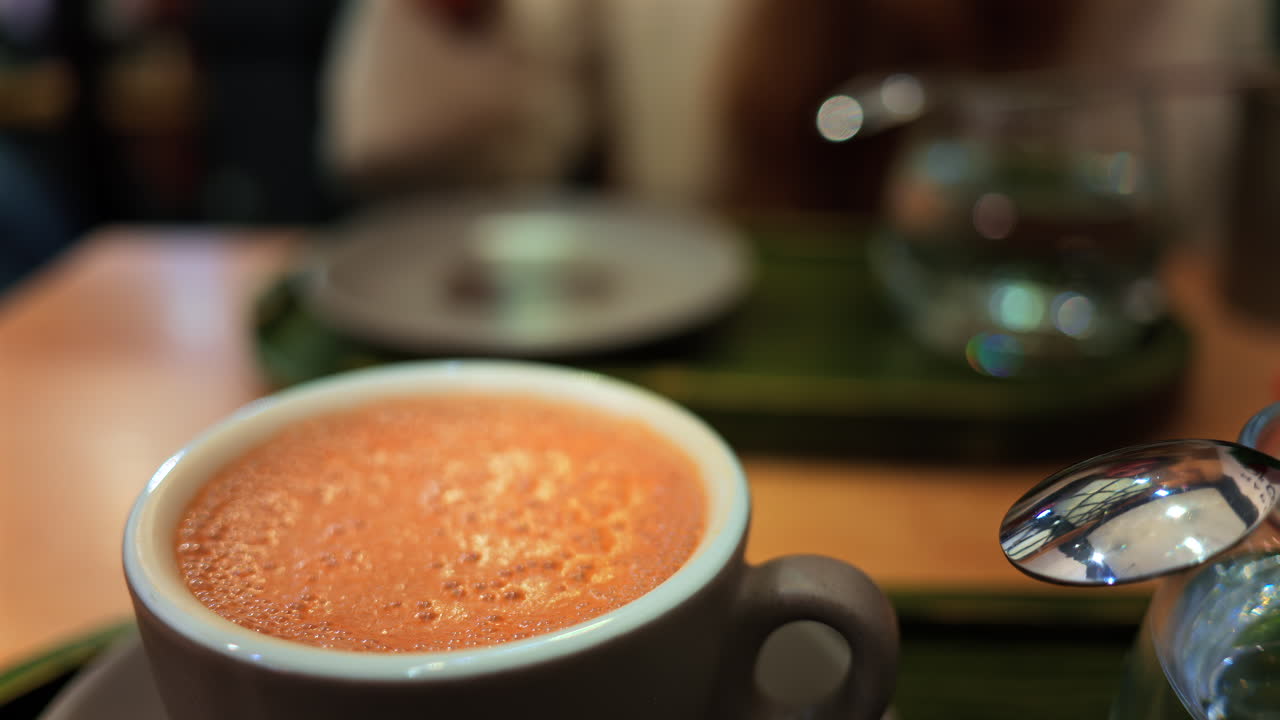 Close up of a cappuccino on a table at a cafe with a blurred view of a woman drinking a matcha latte on the background