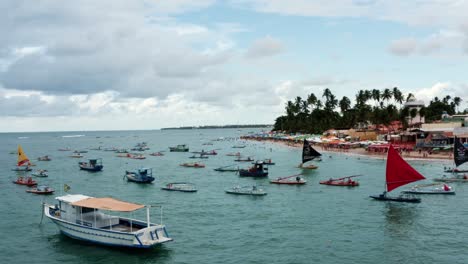 Dolly-out-aerial-drone-shot-of-the-Porto-de-Galinhas-or-Chicken-Port-beach-with-anchored-sailboats,-colorful-umbrellas,-and-tourists-swimming-in-the-crystal-clear-ocean-water-in-Pernambuco,-Brazil