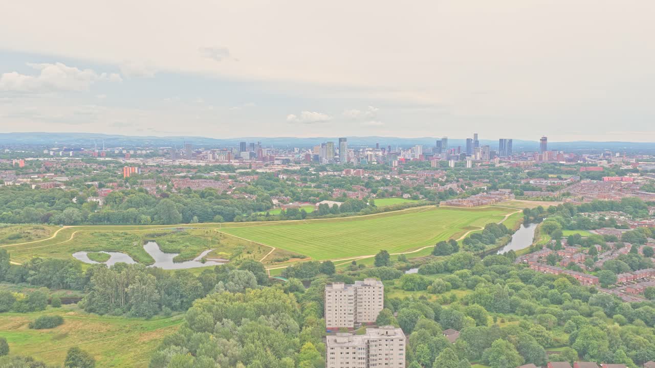 The area near Salford City FC shows residential tower blocks, green parkland, and the Manchester skyline on the horizon, captured in a slow drone glide