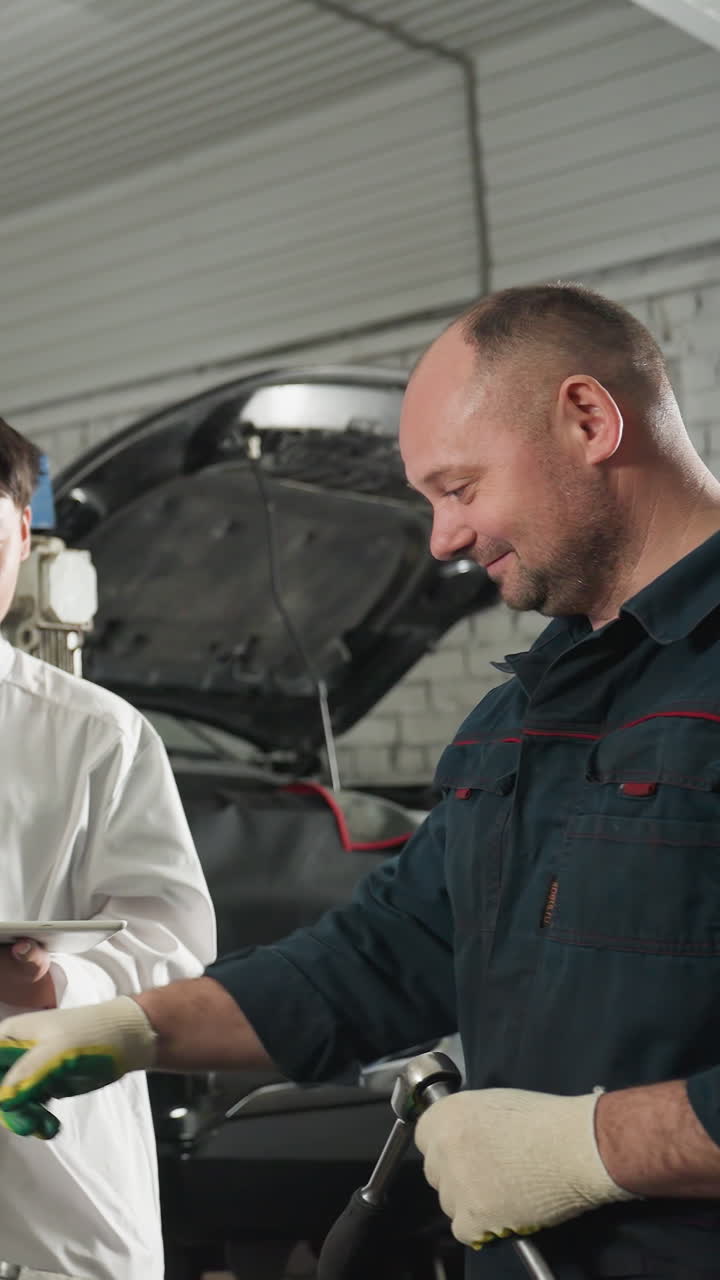 un mecánico demuestra herramientas a los estudiantes mientras una estudiante graba datos en una tableta, un estudiante observa con una sonrisa a un instructor con guantes que levanta el pulgar en un taller automotriz