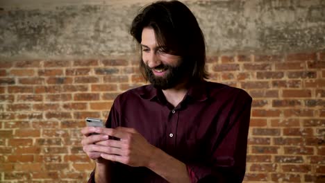 joven moreno agradable con barba está viendo fotos en el teléfono inteligente, riendo, fondo de ladrillo, concepto de comunicación