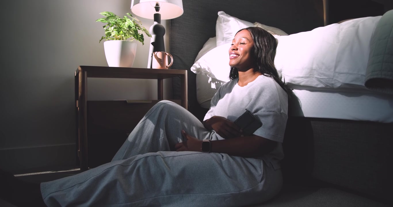 Donna afroamericana seduta in camera da letto, appoggiata a un cuscino, che sorride al sole, prende una tazza e sorseggia.