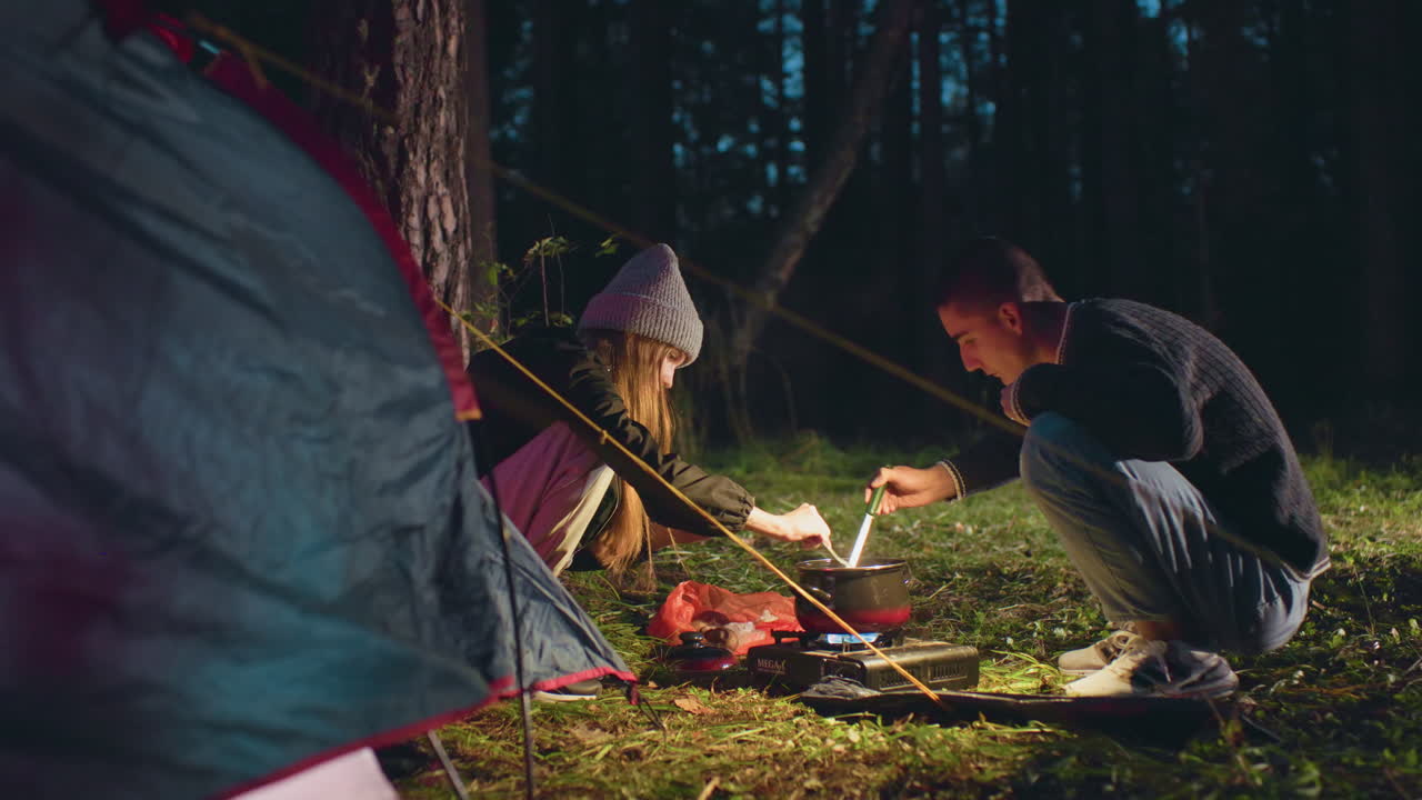 Siblings prepare dinner beside tent during forest camping as girl adds canned food and boy stirs steaming pot using phone light, surrounded by grass and trees under evening sky near portable stove