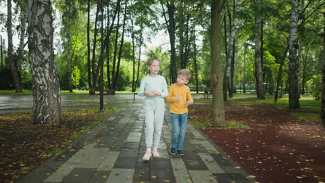Two children stroll along wet paved path surrounded by tall trees in peaceful park, autumn leaves scattered on ground reflect serene outdoor moment in fresh green environment after rainfall