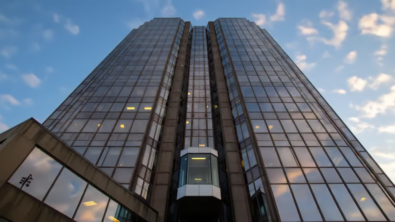 A tall, glass skyscraper stands prominently against the evening sky, reflecting hues of orange and blue as the sun sets. The building's sleek design highlights modern architecture in the city.
