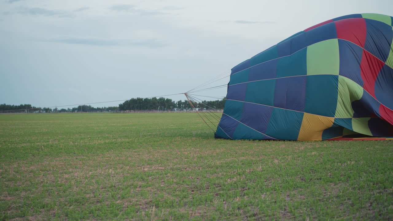 crew members dragging deflated multicolor hot air balloon envelope across green farmland at twilight while guiding tether line and flattening canopy during methodical pack down