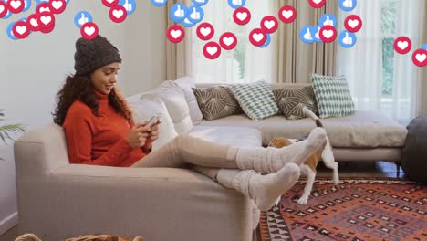 like and heart icons against african american woman using smartphone while her dog on the couch