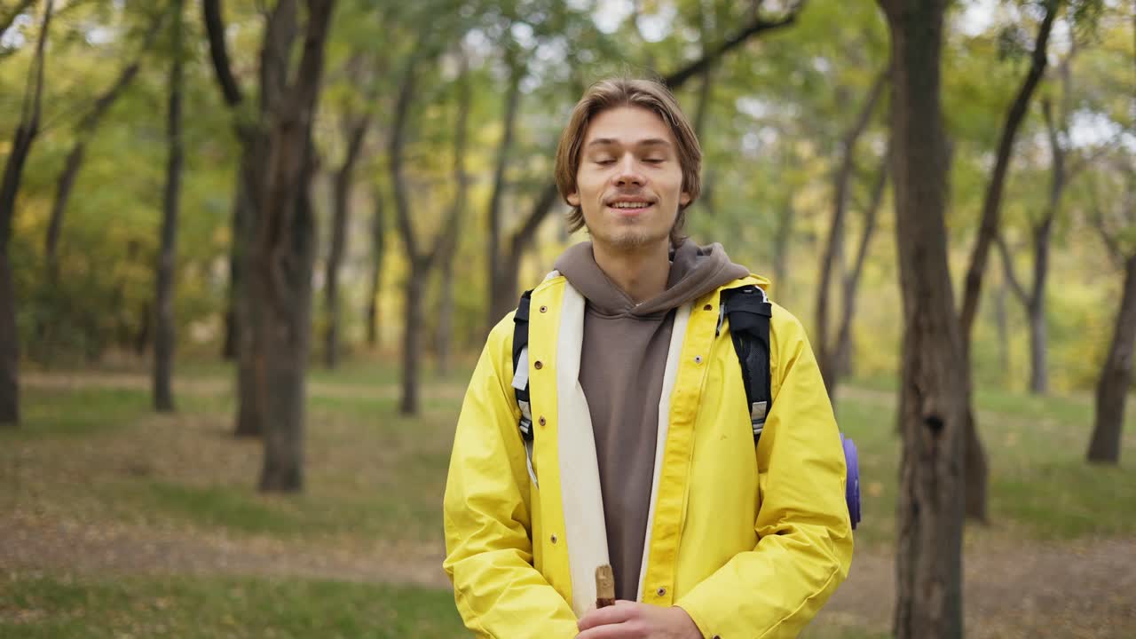 joven alegre con abrigo amarillo con mochila de pie y sonriendo en el bosque