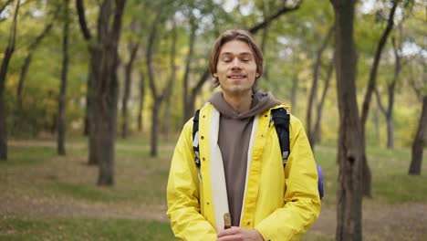 cheerful young man in yellow coat with backpack standing and smiling in forest