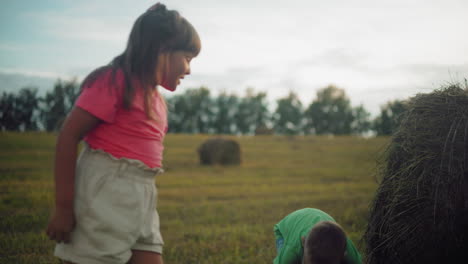 boy in green shirt playfully throws hay at his sister and runs away laughing, the siblings enjoy a carefree moment in a vast countryside field surrounded by hay bales