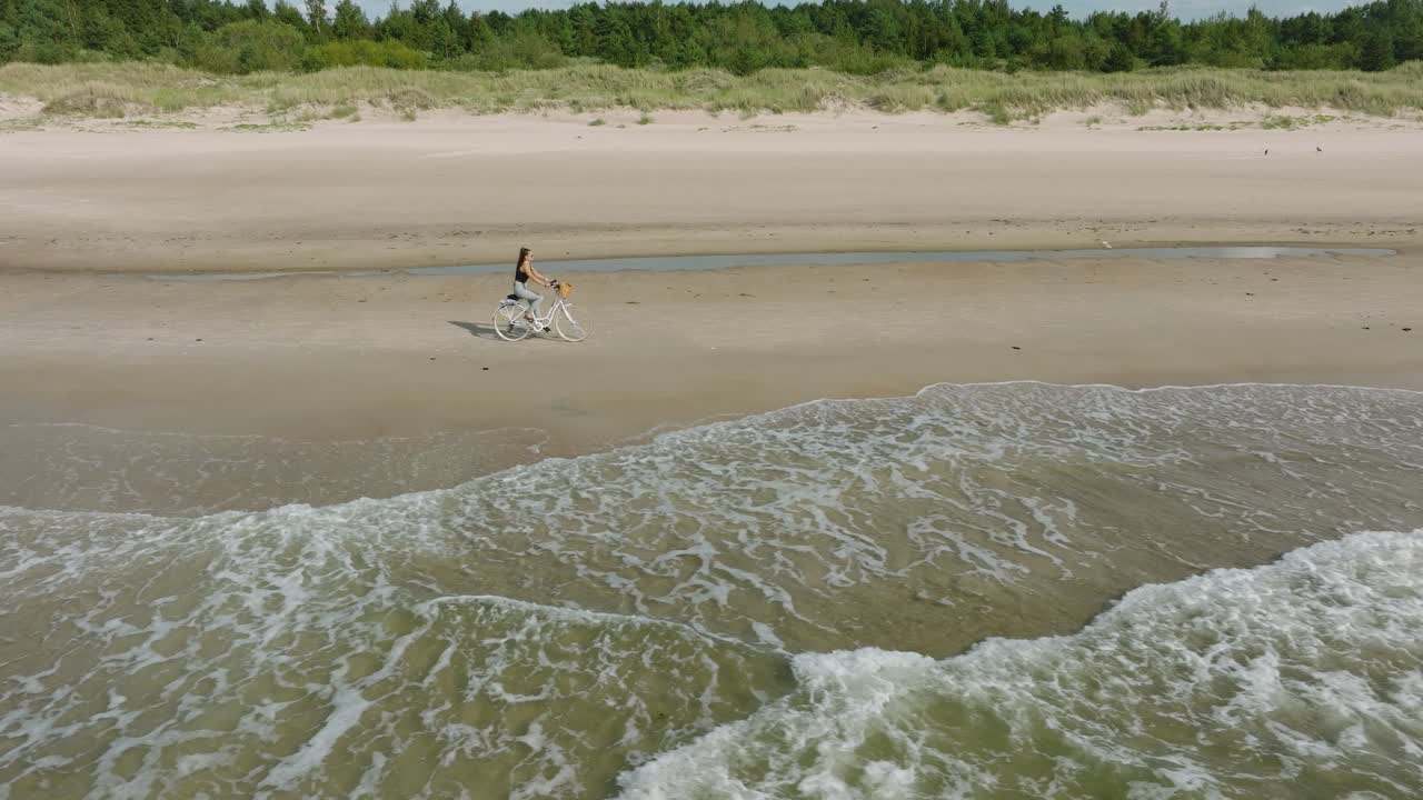 Vista aerea con una giovane ragazza dai capelli lunghi che va in bicicletta sulla spiaggia sabbiosa, giornata di sole, spiaggia di sabbia bianca, concetto di stile di vita attivo, ampia carrellata con drone in movimento verso destra