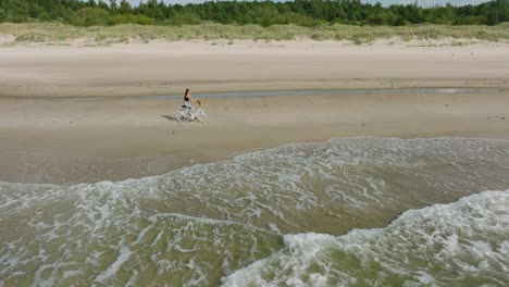 Vista-Aérea-Con-Una-Joven-De-Pelo-Largo-Montando-Una-Bicicleta-En-La-Playa-De-Arena,-Un-Día-Soleado,-Una-Playa-De-Arena-Blanca,-Un-Concepto-De-Estilo-De-Vida-Activo,-Un-Amplio-Tiro-Con-Plataforma-Rodante-Moviéndose-Hacia-La-Derecha