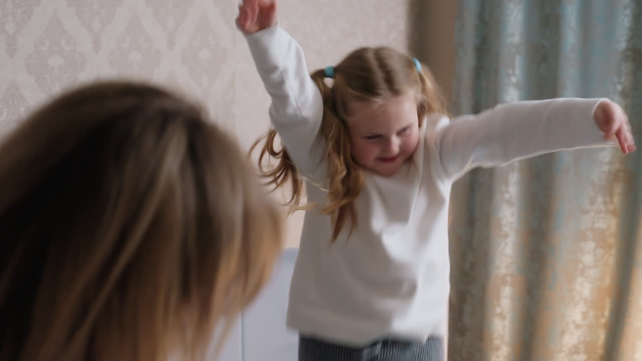 Little girl with pigtails joyfully dances in living room wearing white sweater and gray pants, smiling with excitement while mother watches, capturing playful energy, carefree childhood spirit, and family