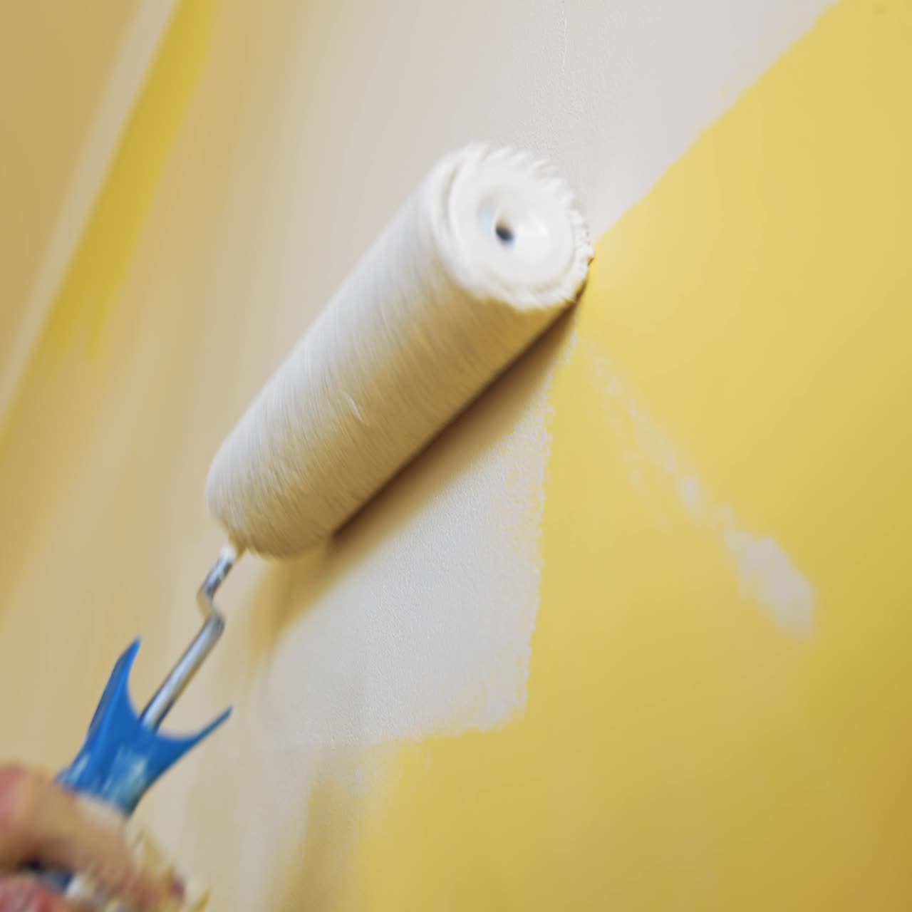 Unrecognizable worker painting wall. Paint roller with light color in man's hand moving up and down on the wall indoors. Room renovation. Close-up