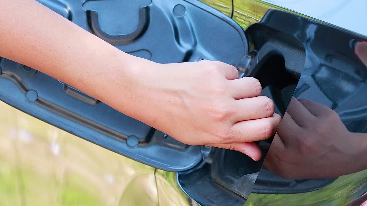 A person's hand opens a vehicle's fuel tank door and unscrews the gas cap