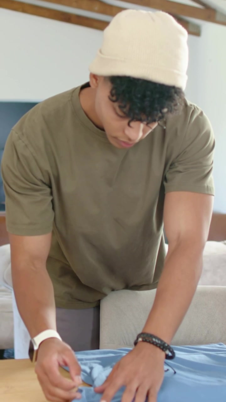 Video vertical: Hombre con camiseta verde oliva viendo arrugas, alisando una camiseta azul en una mesa de madera para aplanarla.