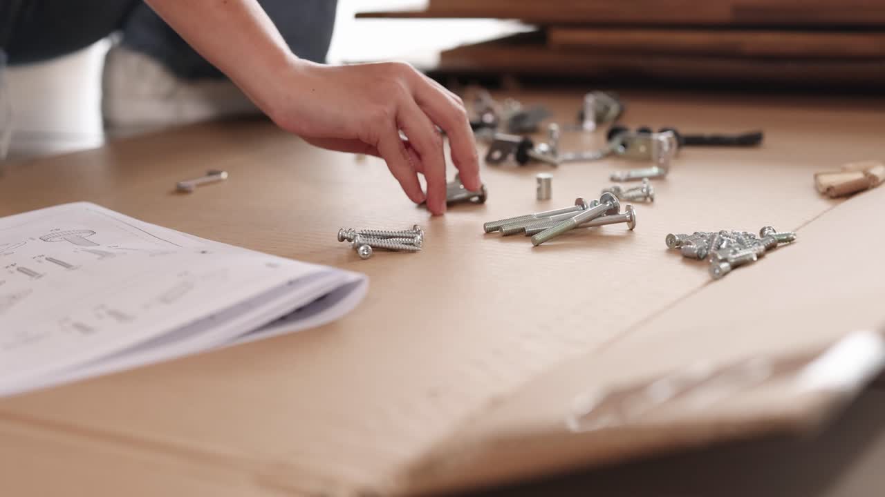 Hands organize hardware on a wooden table with assembly instructions. Bright lighting highlights the detailed process