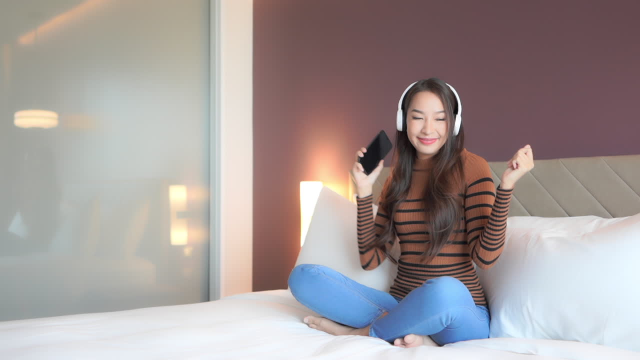 While listening to music through her smartphone and headphone, a young woman is dancing in place on a luxury hotel bed