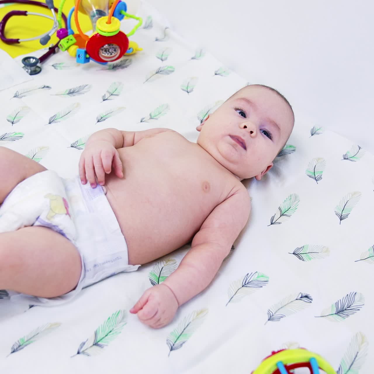 Naked baby in diaper only lying on white sheet. Medical devices are next to the baby. Bright toys around the child