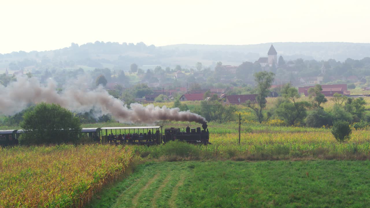 Stunning aerial view captured by a drone of a historic steam train on a nostalgic journey. The locomotive passes fields and a picturesque Romanian village with a fortified church