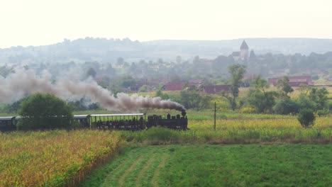 Stunning aerial view captured by a drone of a historic steam train on a nostalgic journey. The locomotive passes fields and a picturesque Romanian village with a fortified church