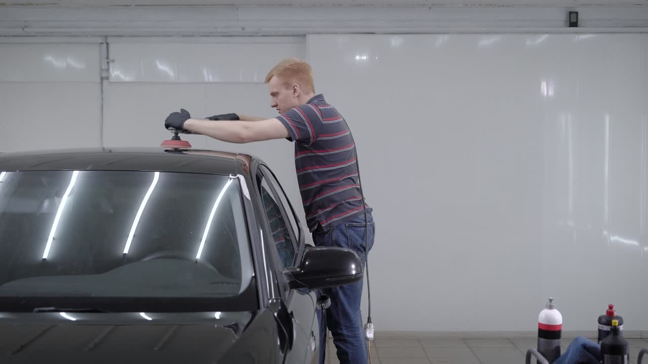 빨간 머리 남자는 차의 표면을 매끄럽게 하기 위해 폴리싱 기계를 사용하고 있으며, 차고의 자동차의 지붕을 도구로 만지고 있습니다.