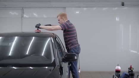 hombre pelirrojo está usando una máquina de pulir para suavizar la superficie del coche, tocando con una herramienta un techo de auto en un garaje