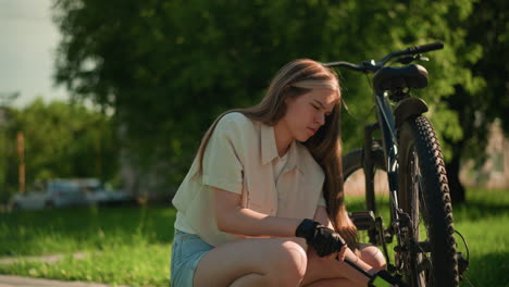 lady in gloves crouched beside bicycle, focused as she inflates back tire on sunlit park path, slightly frustrated, she pauses to check tire's fullness before continuing, surrounded by greenery