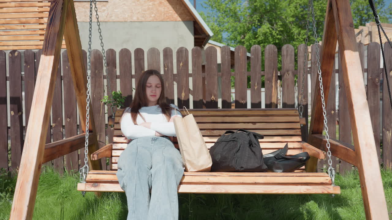 Young lady sits quietly on wooden swing with crossed arms and shopping bags beside her, appearing thoughtful while another person lays on nearby swing in sunny fenced backyard