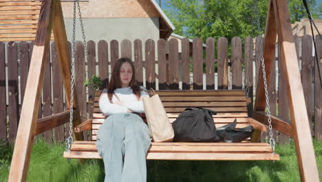 young lady sits quietly on wooden swing with crossed arms and shopping bags beside her, appearing thoughtful while another person lays on nearby swing in sunny fenced backyard