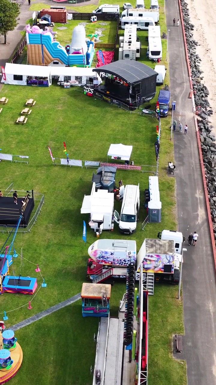 Drone camera glides above vibrant fairground rides, tents, and vehicles along grassy Scottish coastline