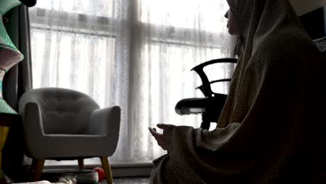 Inside-the-household,-near-a-window-and-leisure-chair,-Muslim-women-pray-while-covered-in-traditional-attire,-embodying-the-practice-of-religious-devotion-at-home