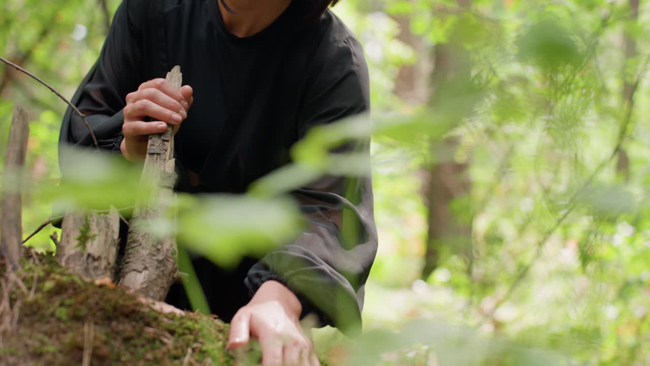 Woman in black flowing dress gently touches moss on fallen tree in lush green forest, sunlight filtering through leaves, creating peaceful and reflective moment filled with texture