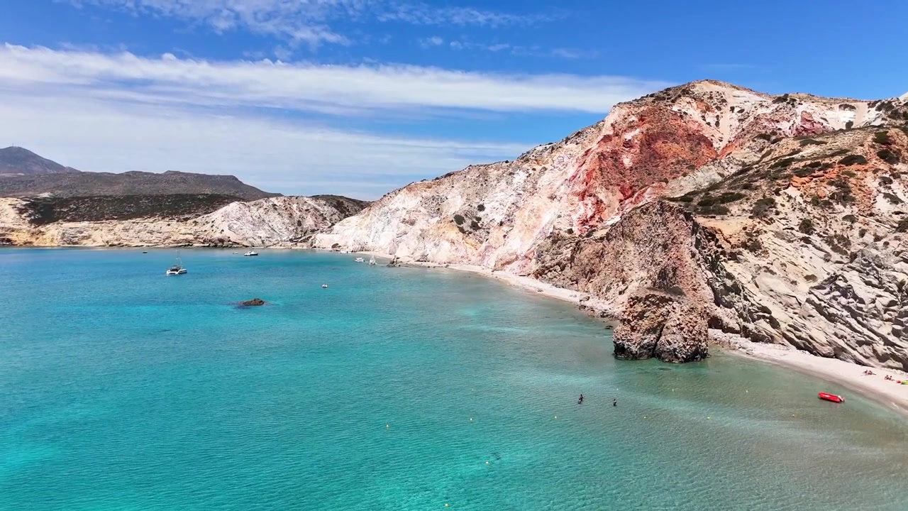 La spiaggia di Firiplaka si trova sotto scogliere vulcaniche rosse, ocra e bianche nel comune di Milo, Milo, Egeo Meridionale, Grecia, con bagnanti lungo la baia riparata, ripresa aerea con drone