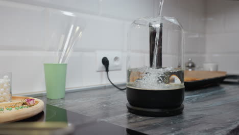 el agua se vierte en un frasco de vidrio en un mostrador de la cocina. el fondo borroso presenta un enchufe de pared, una bandeja con productos horneados, una taza y cuentas decorativas