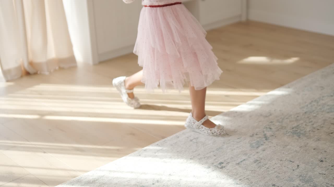 молодая девушка весело танцует в светлой, уютной гостиной. она носит розовое платье с дизайном сердца, играюще движется в солнечном, минималистском пространстве с нейтральным декором и мягким естественным светом.