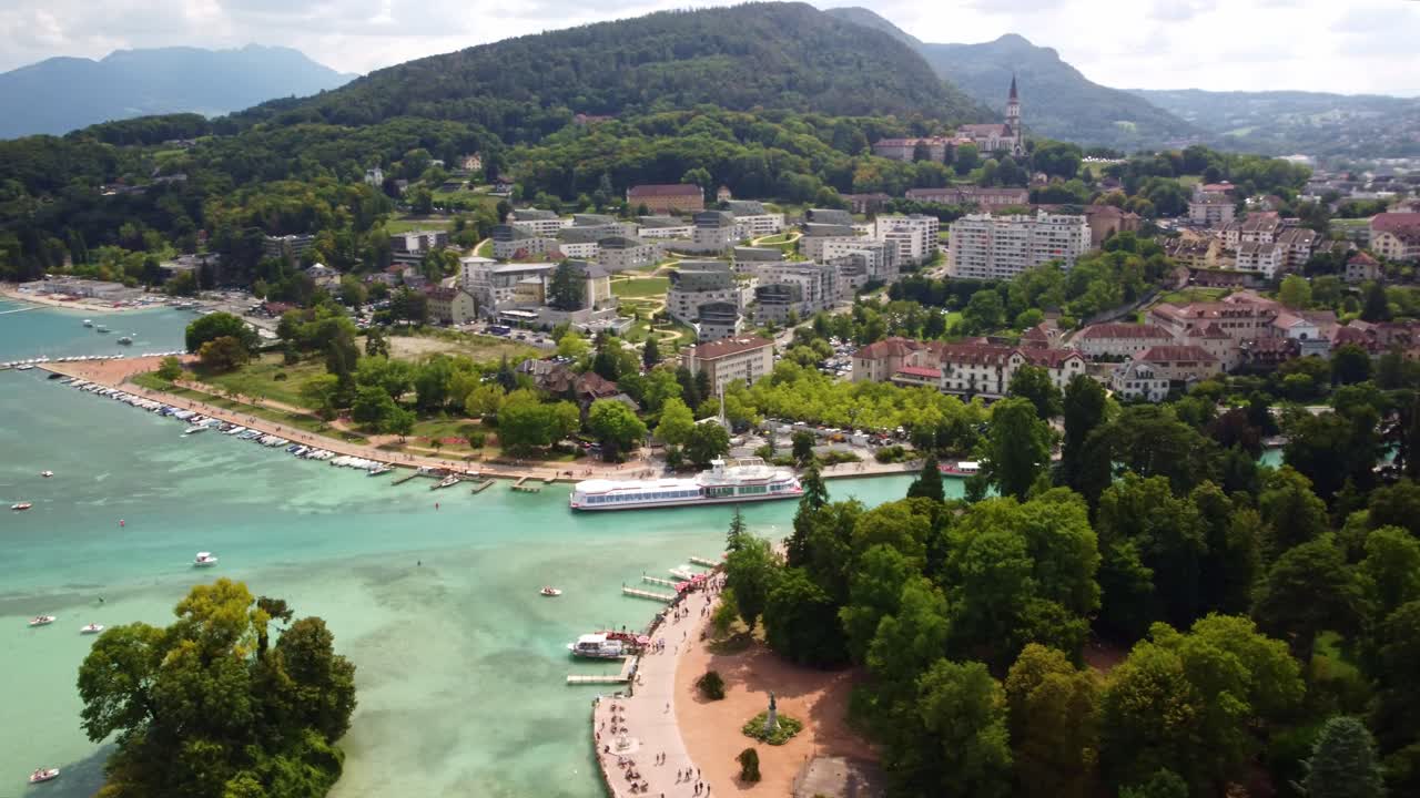 Drone footage rotates around a ferry moored up on River Le Thiou in Annecy. As the footage continues it reveals the stunning views on the city centre. Trees surrounding and hills in the background