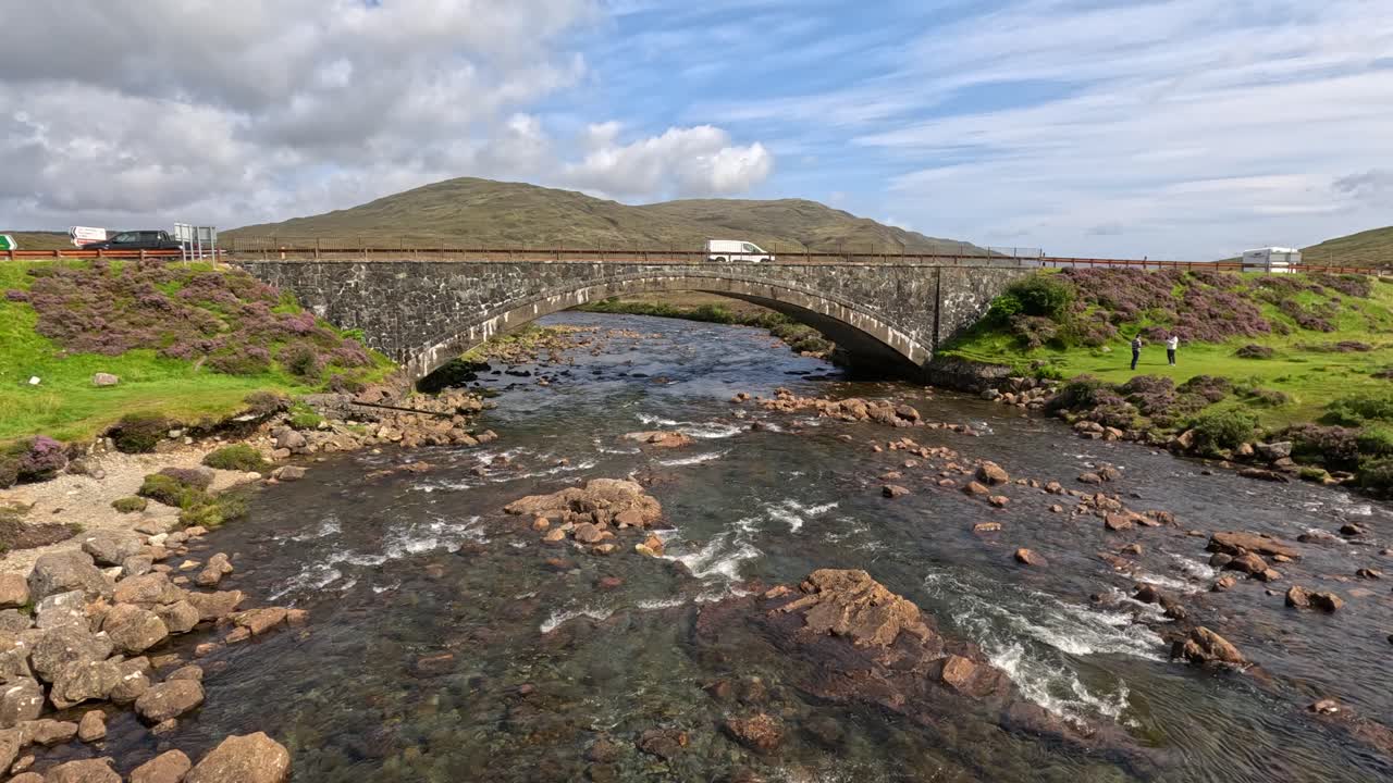 A wide shot captures a stone bridge spanning a clear mountain stream, with vehicles crossing and lush green hills under bright daylight and partly cloudy skies