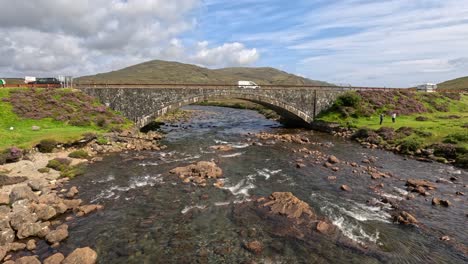 a wide shot captures a stone bridge spanning a clear mountain stream, with vehicles crossing and lush green hills under bright daylight and partly cloudy skies