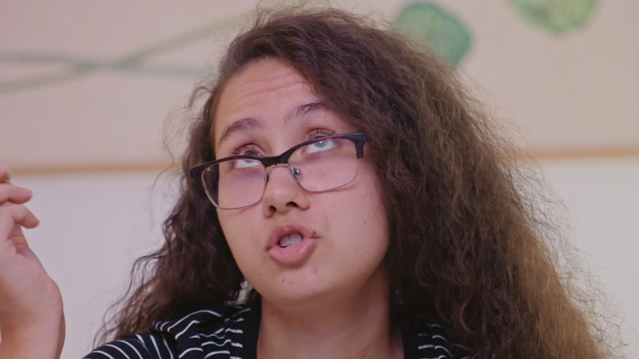 Young woman hotel guest tasting snack while seated in cozy hotel room, wearing glasses and curly hair, looking upward and reacting after catching small treat, candid closeup capturing natural