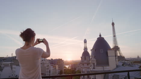 mujer feliz usando un teléfono inteligente tomando fotos disfrutando de compartir la experiencia de las vacaciones de verano en parís fotografiando la hermosa vista del atardecer de la torre eiffel en el balcón