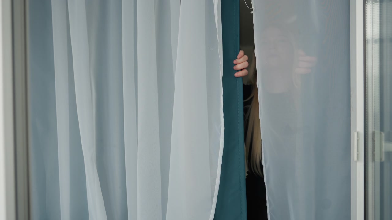Blick durch einen transparenten Vorhang in einer neuen Wohnung. Weicher Stoff verbirgt die Silhouette einer blonden Mieterin während des Umzugstages. Morgenlicht filtert durch. Balkonkästen und kahle Möbel sind sichtbar. Ruhige Atmosphäre.