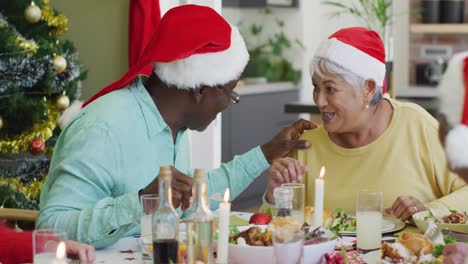 Fröhliches,-Vielfältiges-älteres-Paar-Mit-Weihnachtsmützen,-Das-Sich-Zu-Hause-Am-Weihnachtstisch-Unterhält