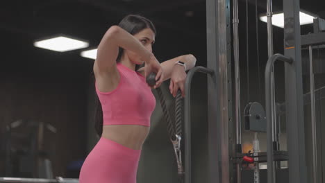 A-hispanic-brunette-woman-in-a-pink-suit-pulls-a-rope-in-a-crossover-with-her-hands-to-train-her-shoulders.-Shoulder-workout-in-a-trainer.-Professional-woman-instructor