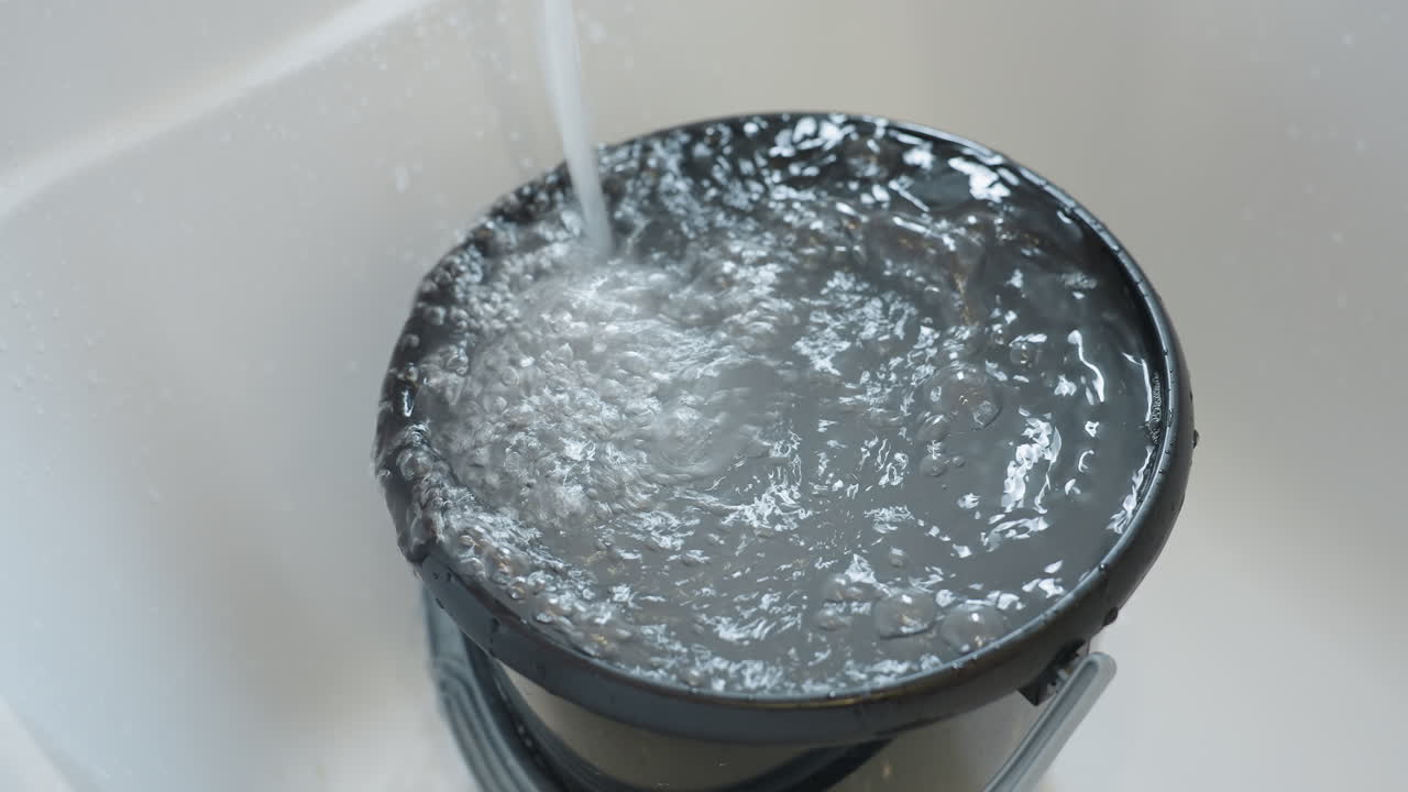 Close up of running water filling black bucket placed in bathtub, surface bubbling and splashing as container overflows, representing household cleaning, sanitation